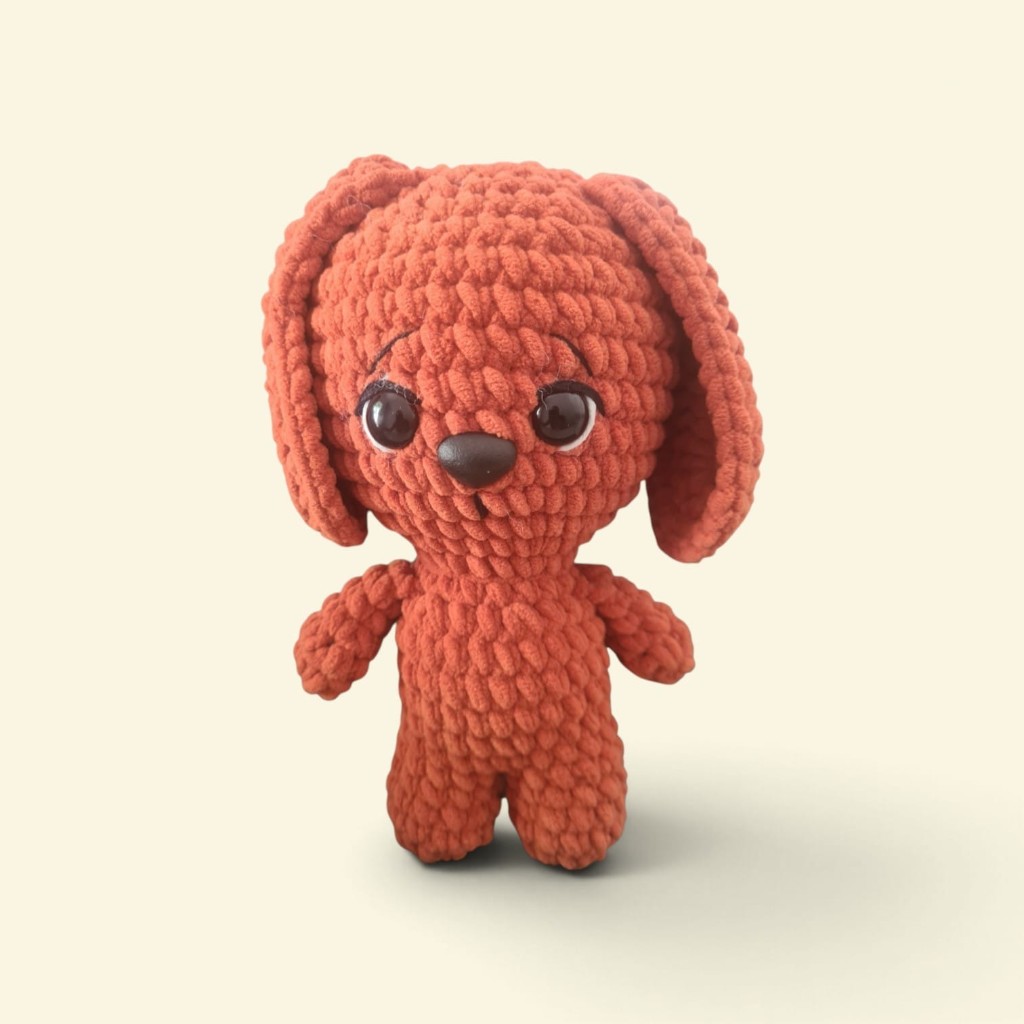 Amigurumi de cachorro em laranja de crochê, visto de frente, com orelhas compridas e focinho marrom.