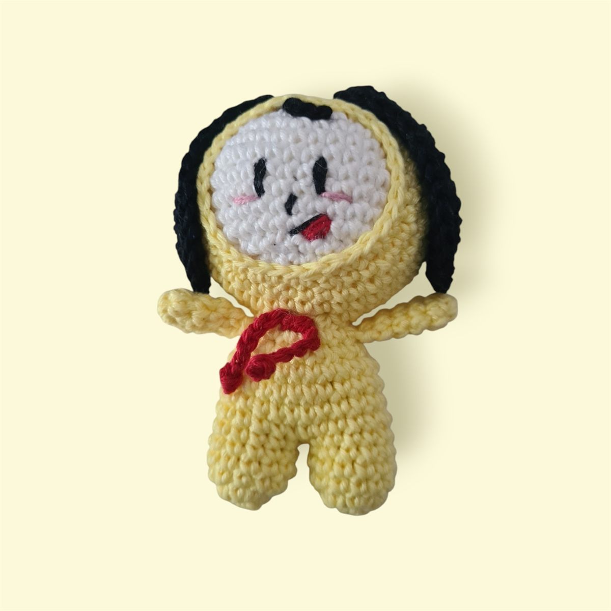 Amigurumi de crochê amarelo com capuz preto, rosto branco e lacinho vermelho.