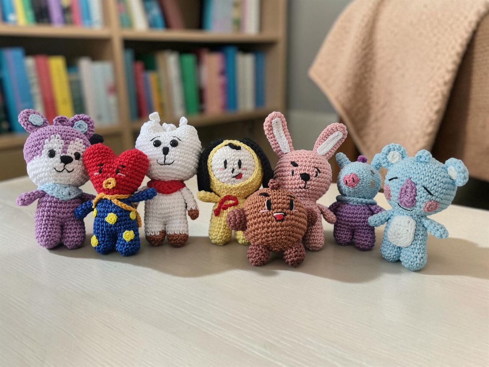Amigurumis de crochê enfileirados em uma mesa, com uma estante de livros ao fundo.