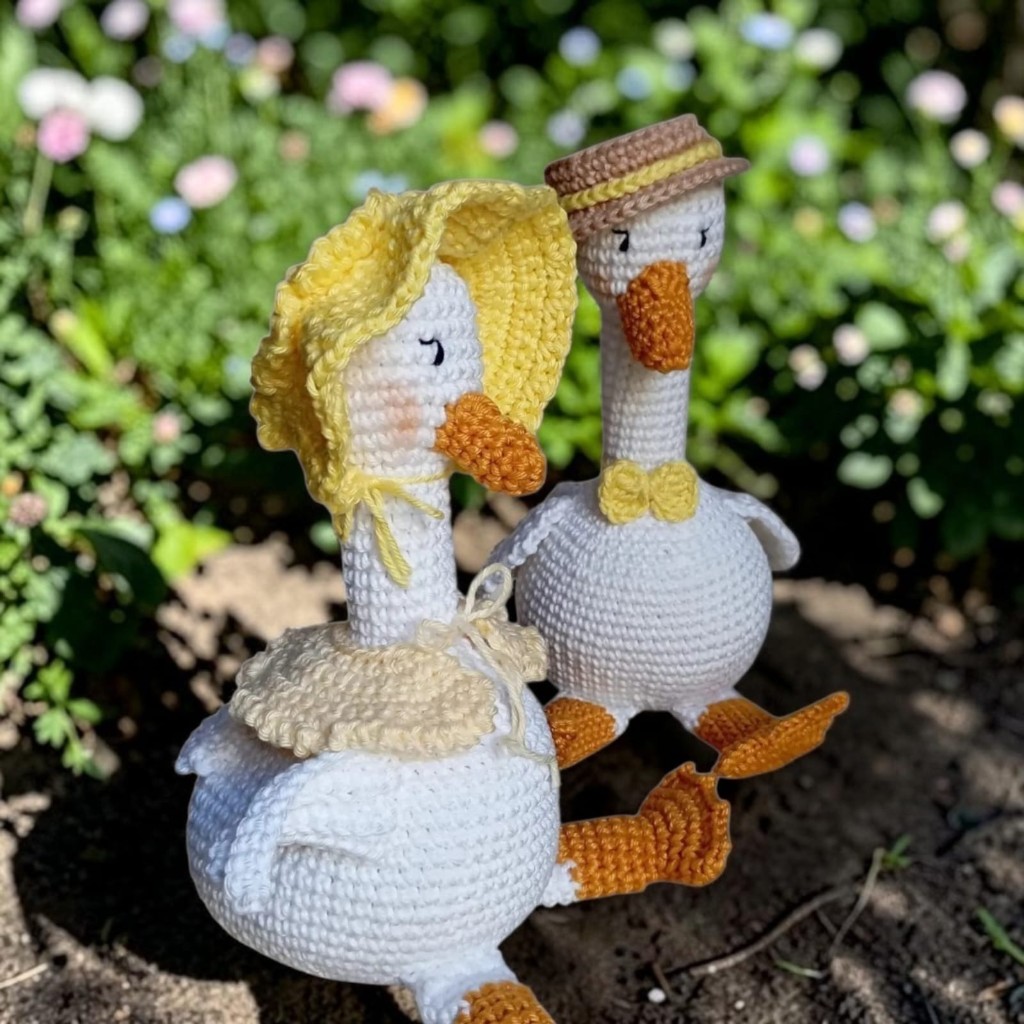Casal de gansos de crochê brancos no jardim. Ela usa chapéu amarelo e ele usa chapéu marrom com gravata borboleta amarela.