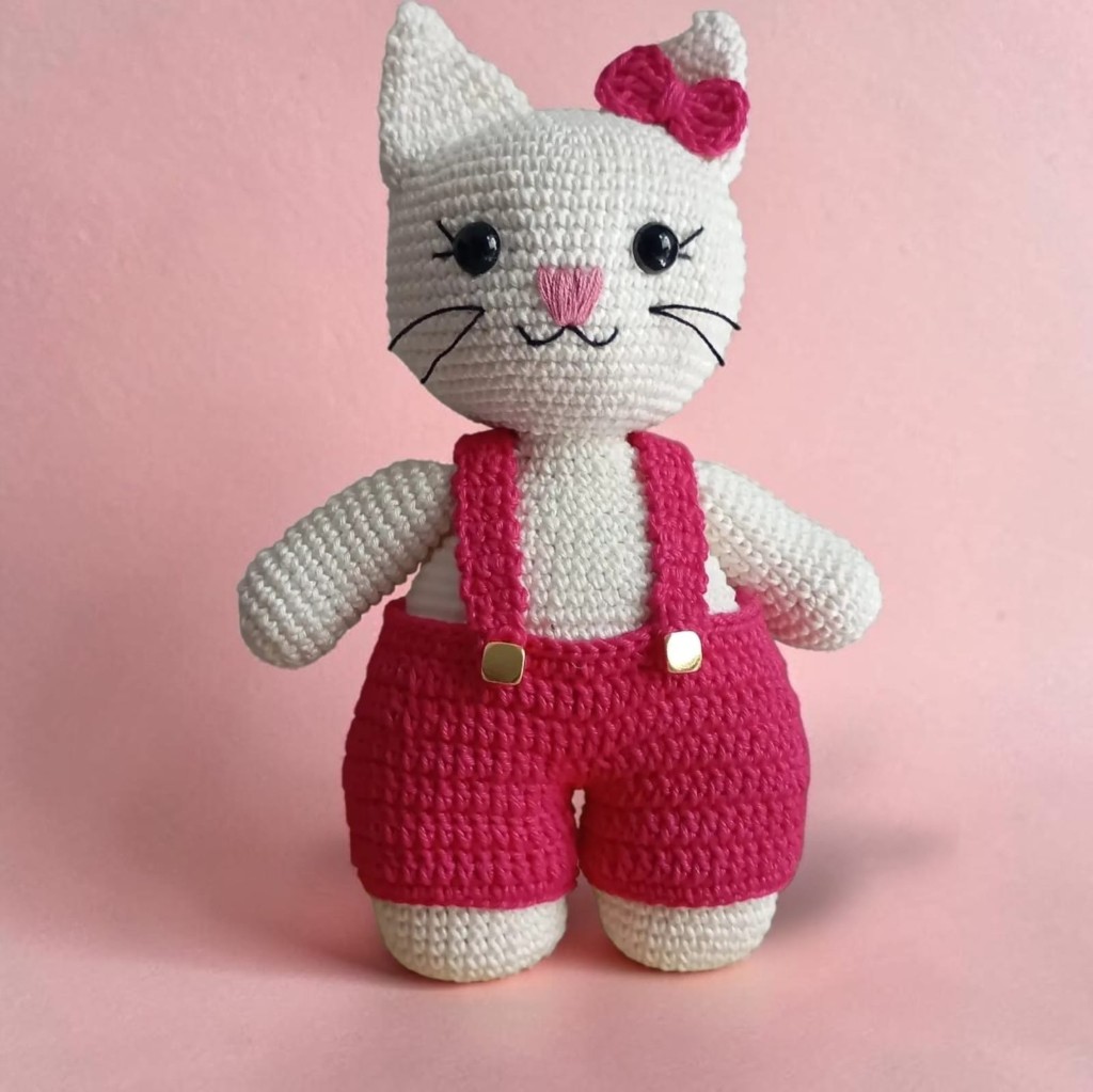 Amigurumi de gatinha branca de crochê, com laço rosa e jardineira pink.