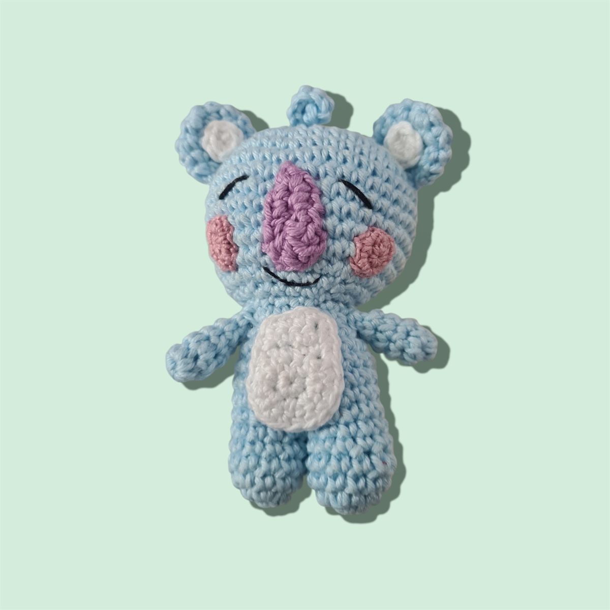 Amigurumi de coala azul de crochê, com focinho lilás, bochechas rosadas e barriga branca.