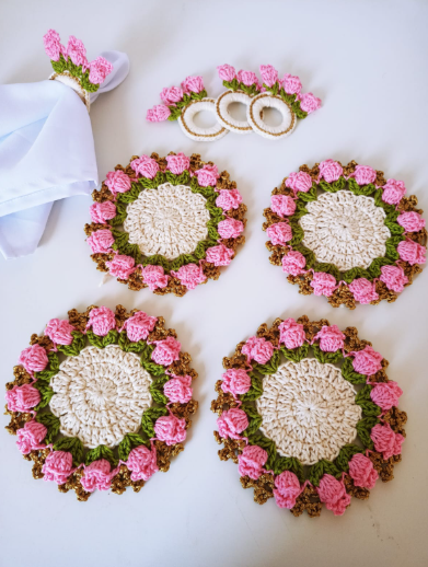Jogo de sousplats de crochê com flores rosa, folhas verdes, centro creme e borda dourada, com porta-guardanapos combinando.