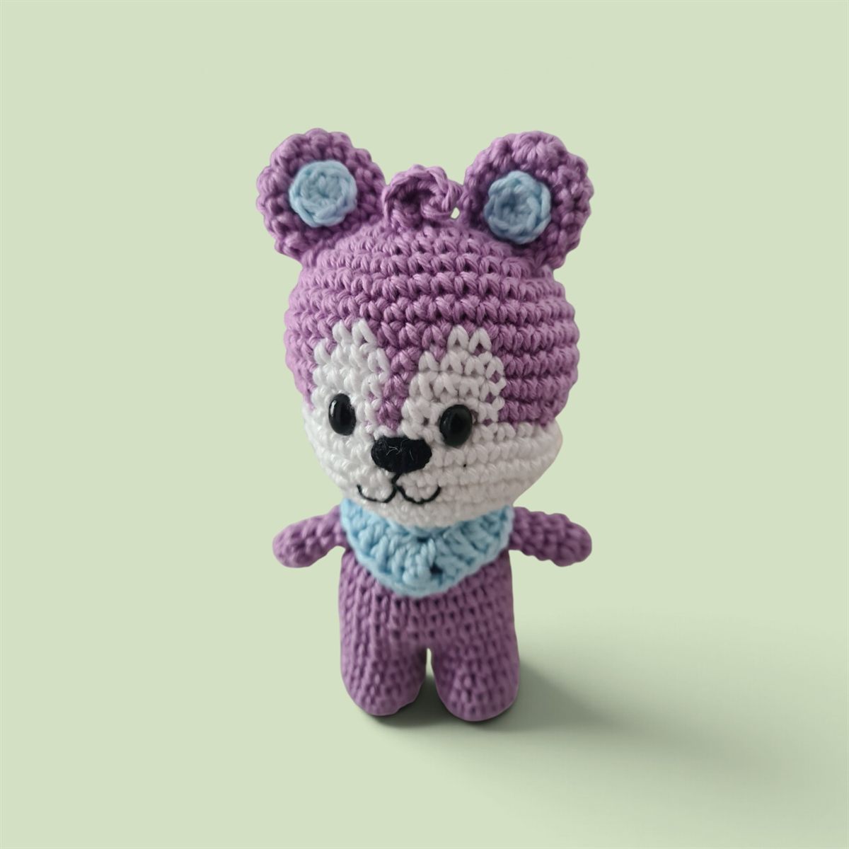 Amigurumi de ursinho lilás de crochê, com focinho branco, orelhas azul-claras e lenço azul.